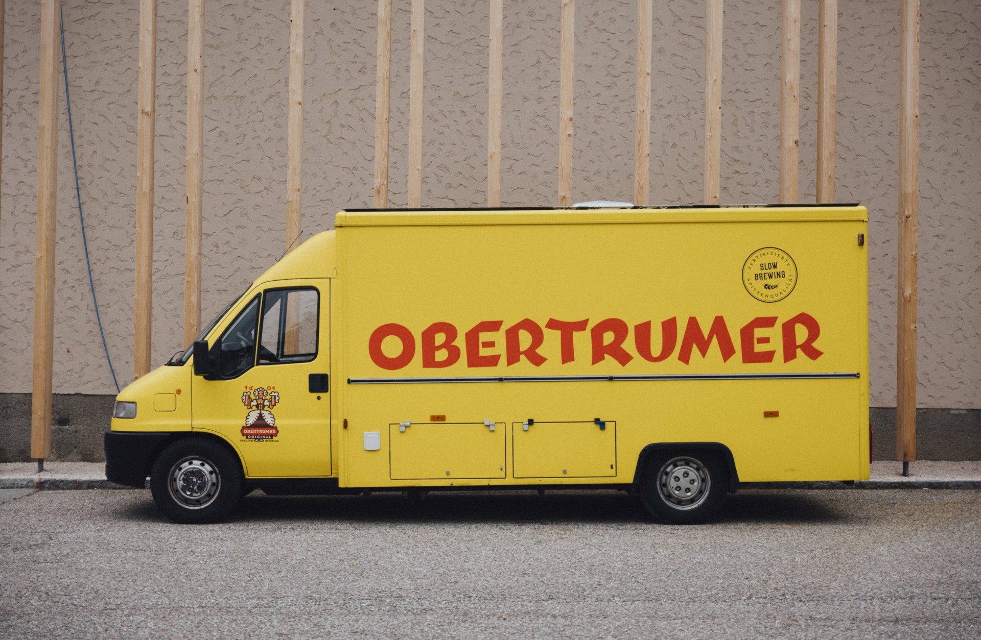 Ein gelber Obertrumer Brauereiwagen parkt vor einer Wand mit Holzdetails. Auf der Seite steht „OBERTRUMER“.