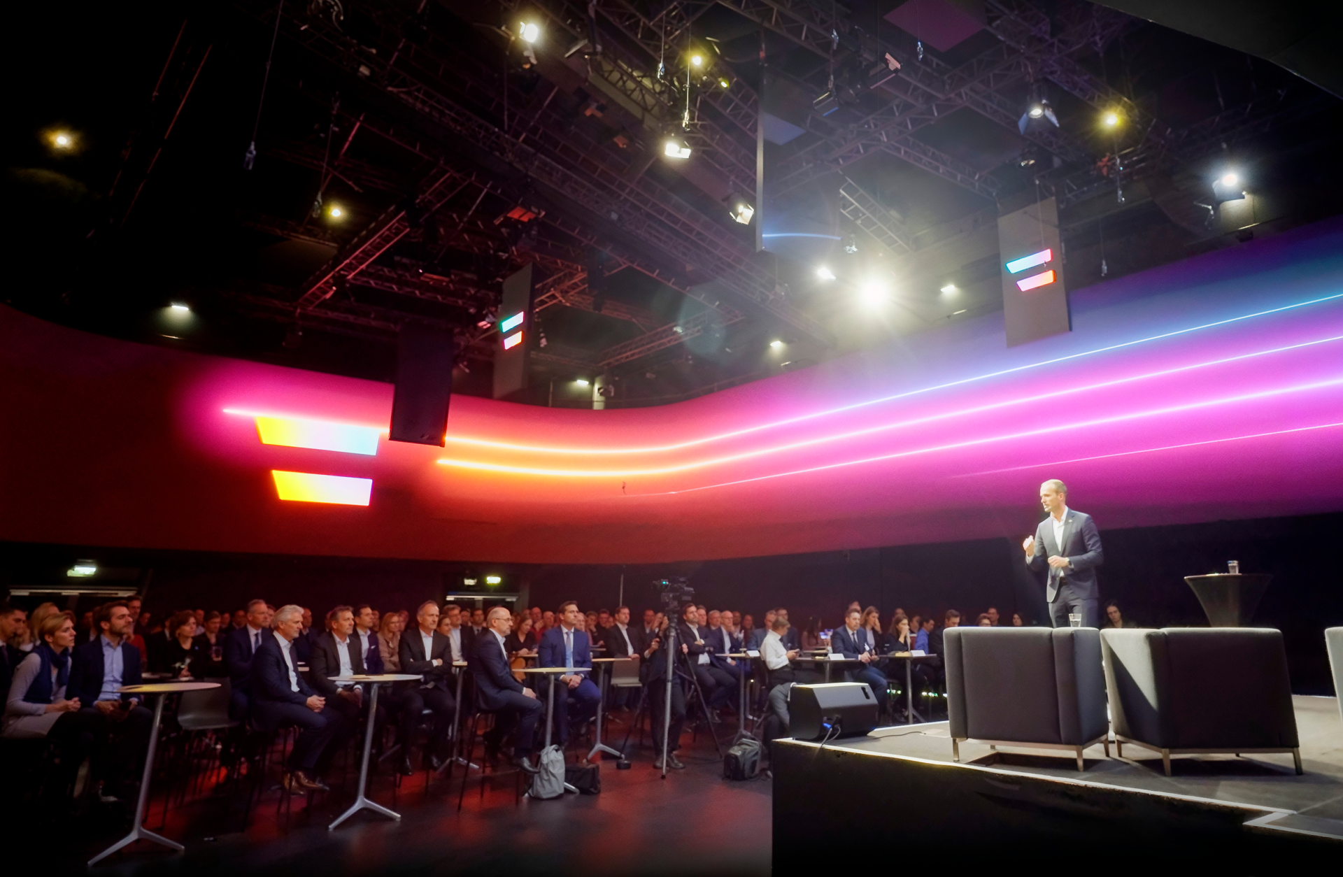 Ein Mann spricht auf einer Bühne zu einem Publikum. Helle Lichtlinien und ein Logo sind im Hintergrund.