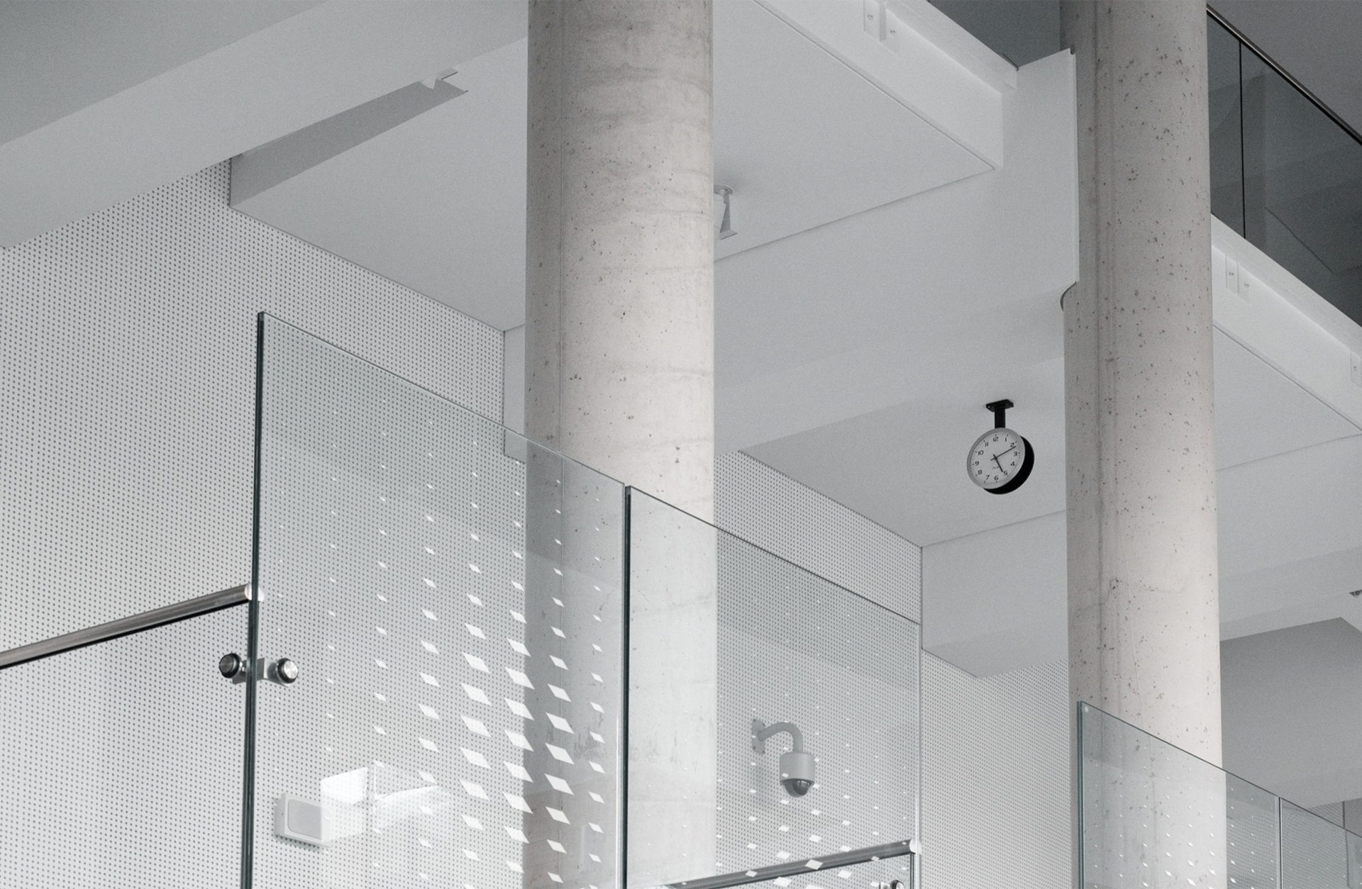 Innenraum mit Betonsäulen, Glasgeländern und einer Wand mit Punktmuster. Eine Uhr hängt an der Decke.