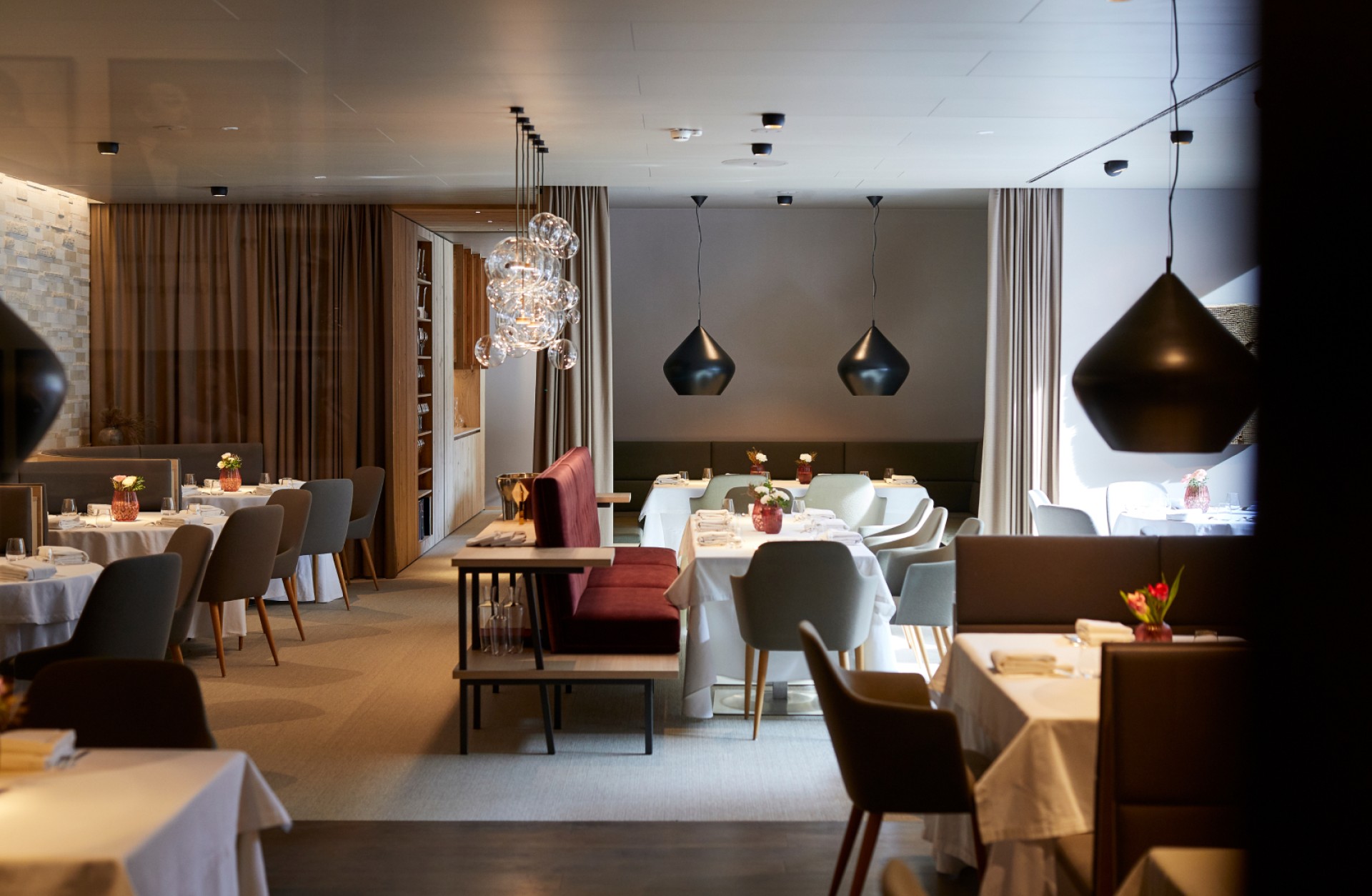 Ein modernes Restaurant mit Tischen, Stühlen, Sofas, Deckenleuchten und Vorhängen in gedämpftem Licht.