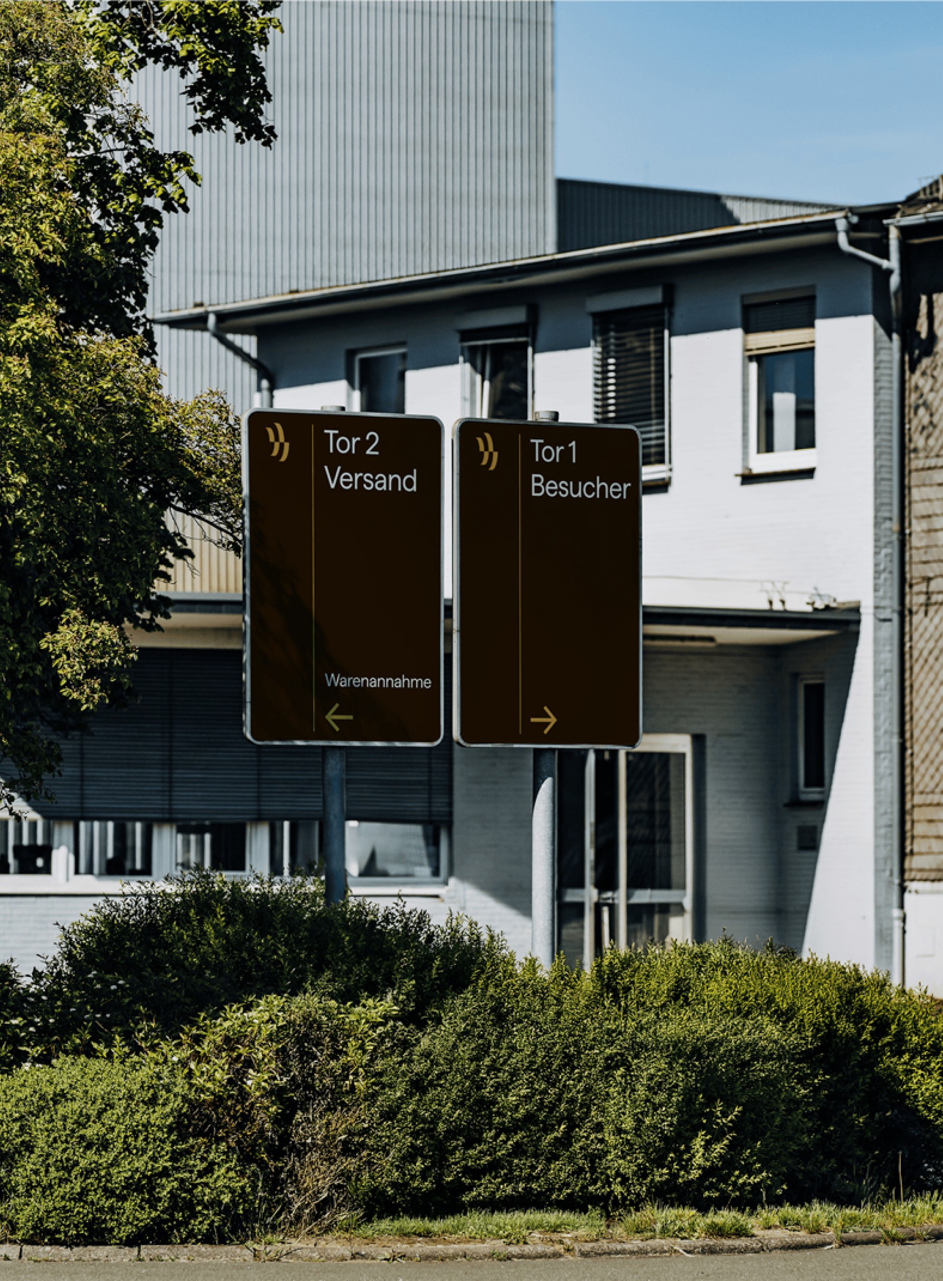 Braune Schilder "Tor 2 Versand Warenannahme" und "Tor 1 Besucher" mit Pfeilen vor einem Gebäude.
