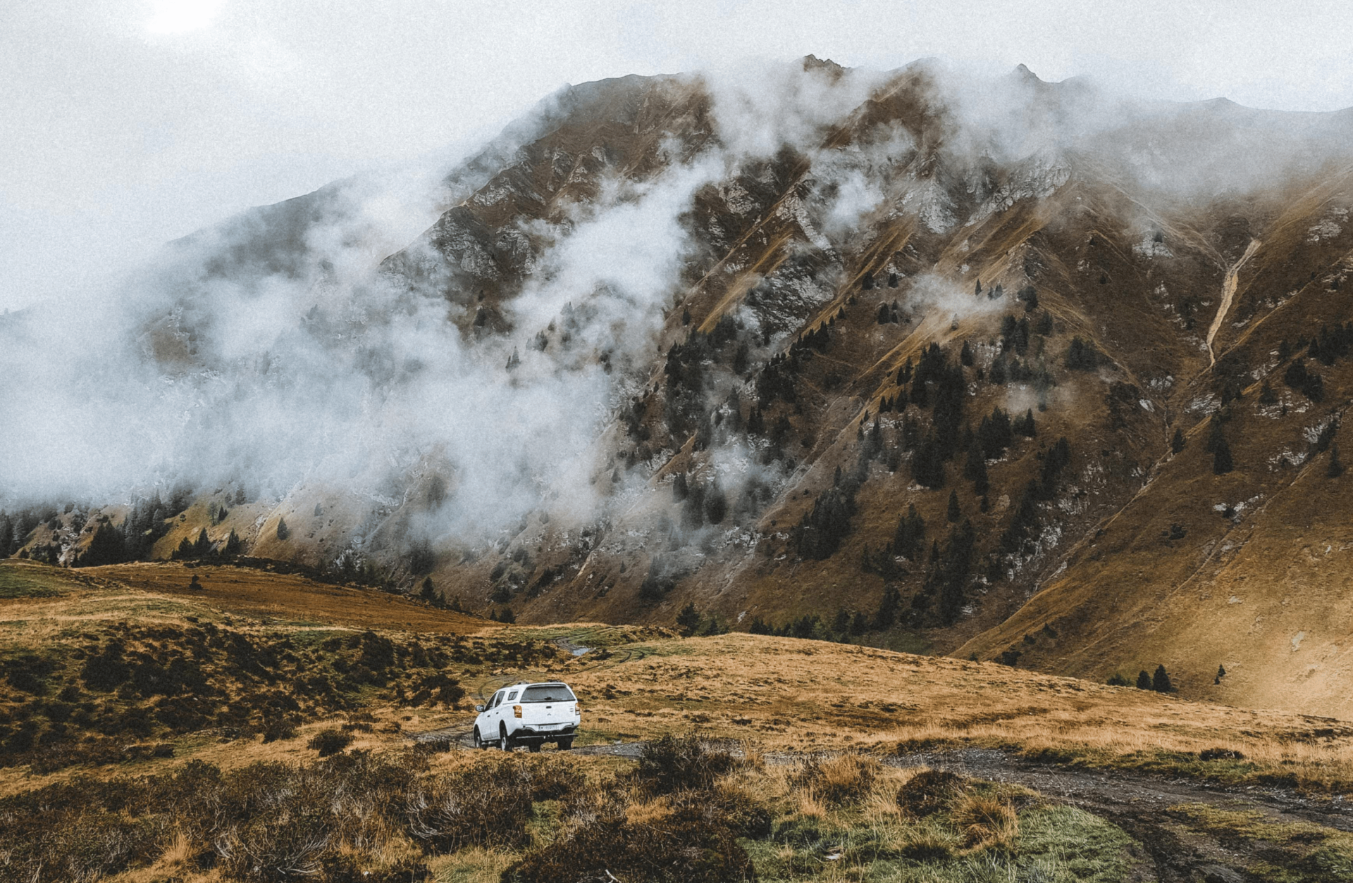 Ein weißer Geländewagen fährt auf einer Straße vor einem nebligen Berg in den Alpen.