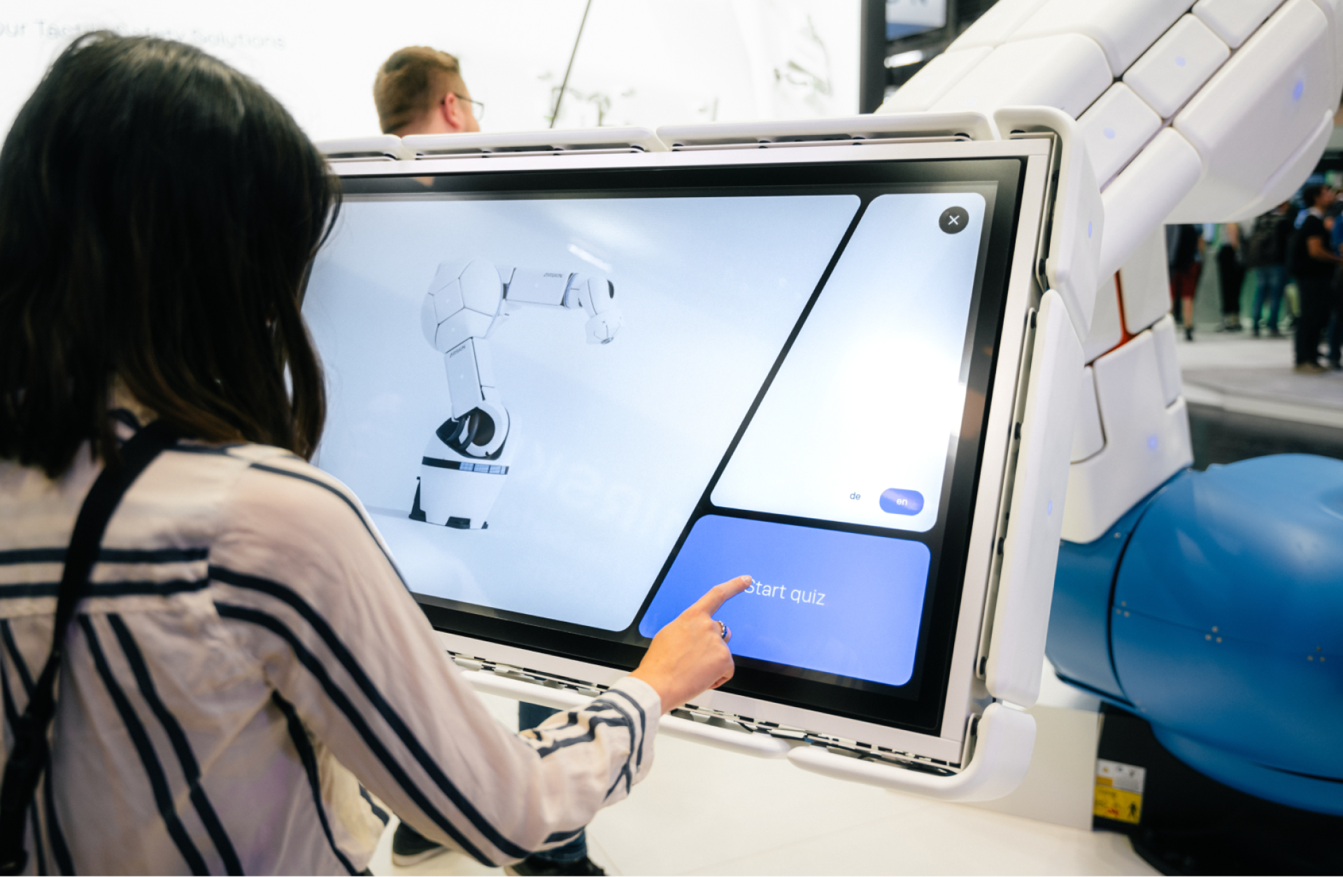 Frau nutzt Touchscreen an Roboterarm. Auf dem Bildschirm ist ein Quiz mit dem Text "Start Quiz".